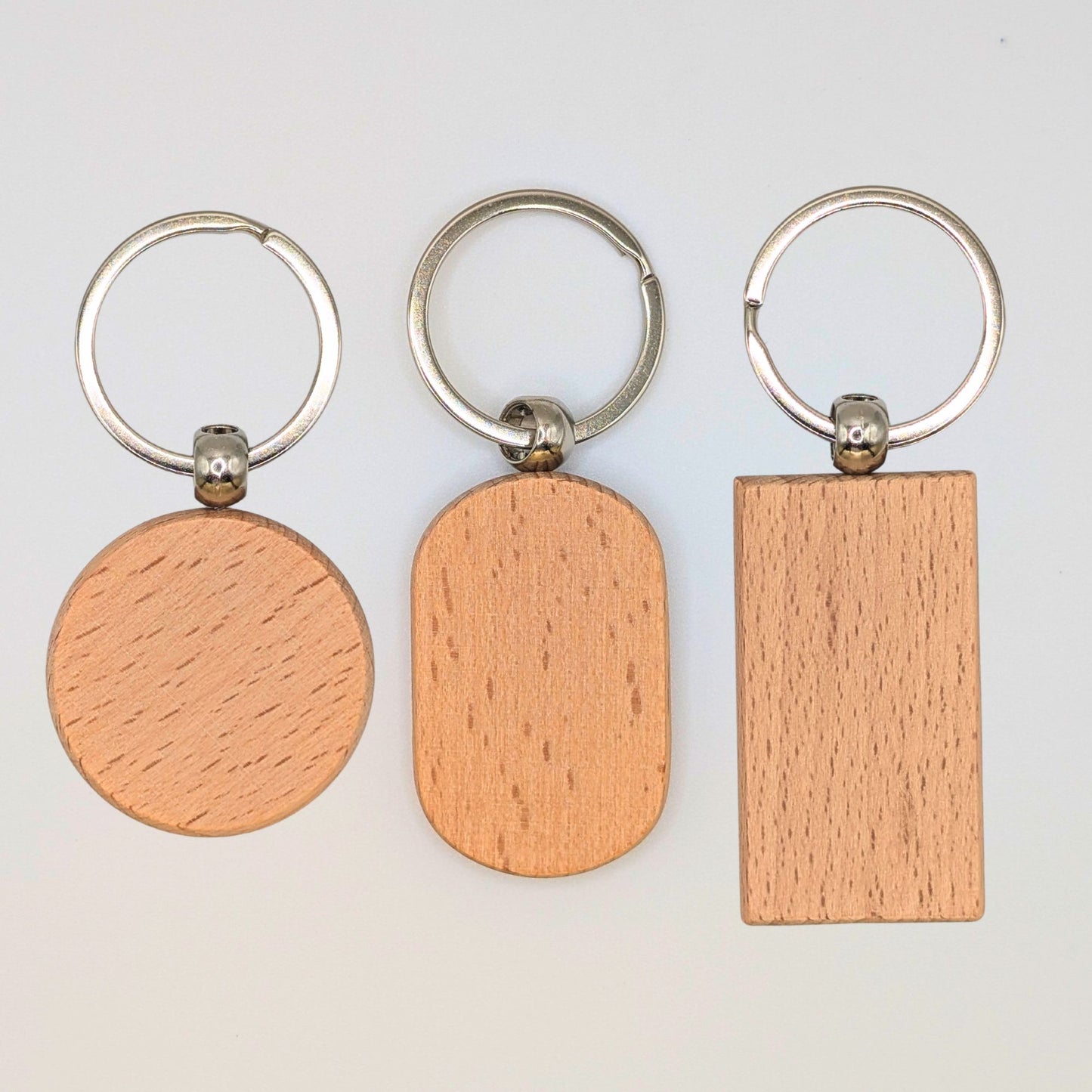Three wooden keychains with silver metal keyrings, shown on a plain light background. The wooden tags have three different shapes: a circle, an oval, and a rectangle, all displaying a light natural wood grain texture.