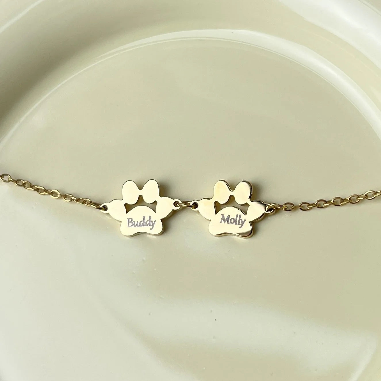 A gold chain bracelet with two paw print charms, one engraved with the name 'Buddy' and the other with 'Molly', displayed on a light beige ceramic surface.