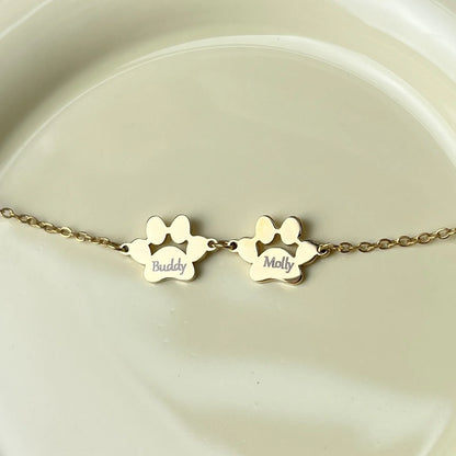 A gold chain bracelet with two paw print charms, one engraved with the name 'Buddy' and the other with 'Molly', displayed on a light beige ceramic surface.