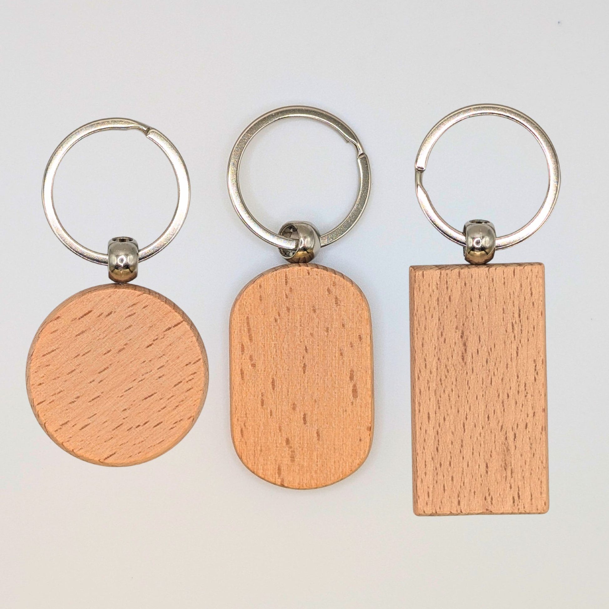 Three wooden keychains with silver metal keyrings, shown on a plain light background. The wooden tags have three different shapes: a circle, an oval, and a rectangle, all displaying a light natural wood grain texture.