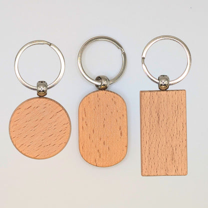 Three wooden keychains with silver metal keyrings, shown on a plain light background. The wooden tags have three different shapes: a circle, an oval, and a rectangle, all displaying a light natural wood grain texture.